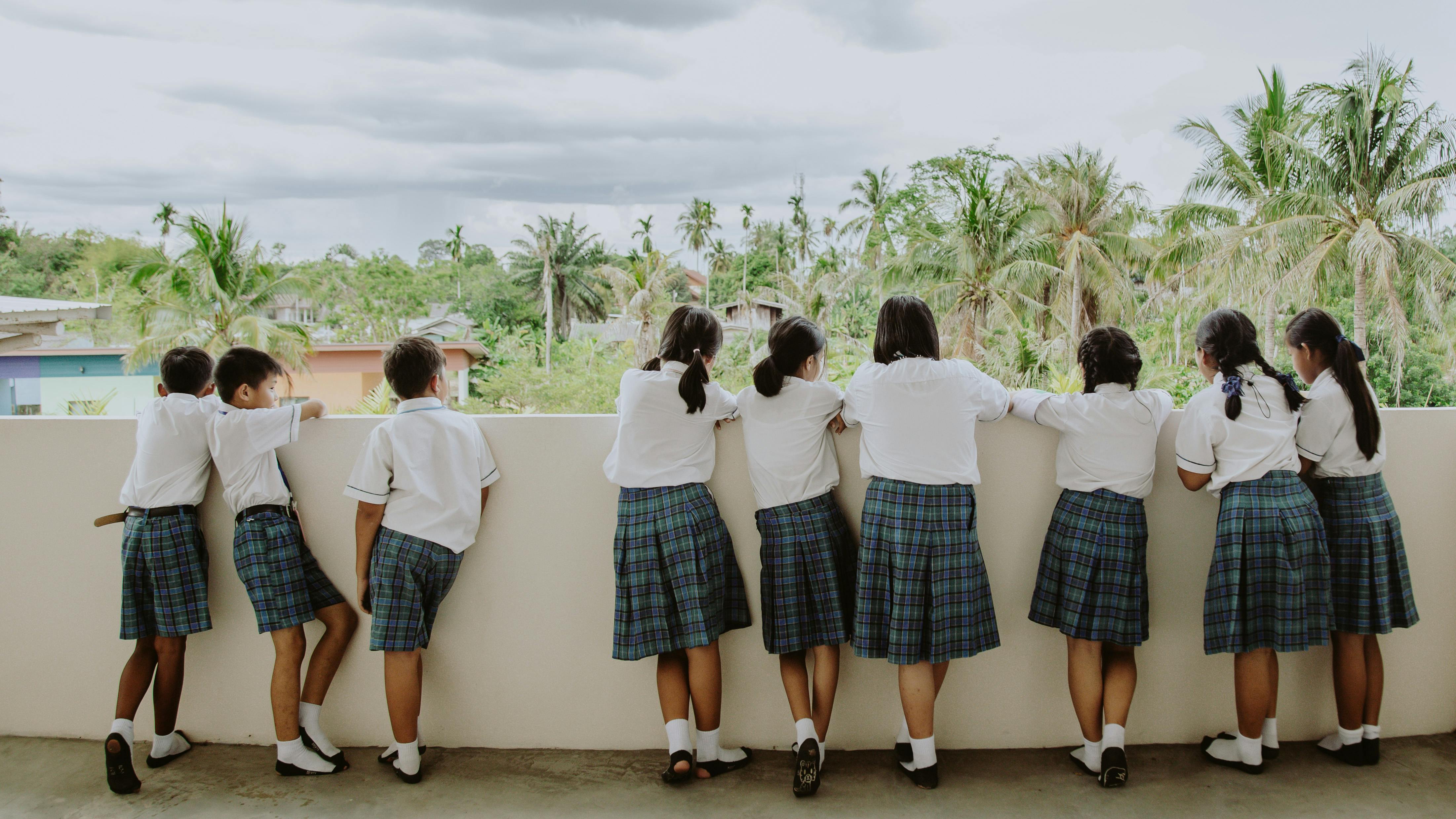 Many school students standing on what seems to be a balcony. They are looking off into the distance with their backs facing us. The background features many trees. The students uniforms are white shirts, and blue, black, white and green plaid skirts.