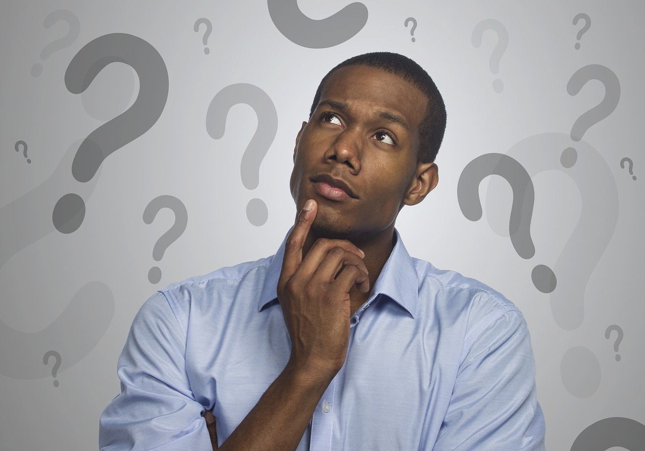 An adult man wearing a blue button up shirt with his pointer finger on his chin. He appears to be deep in thought. Behind him is a grey background that becomes a brighter white directly surrounding the man. On the background are many different question marks in varying sides and shades of grey.