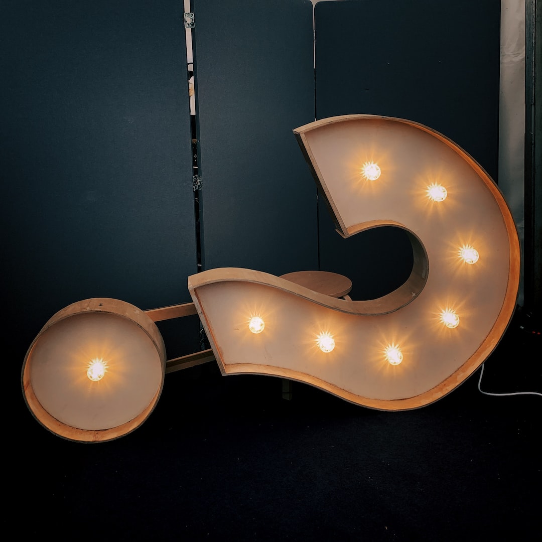 This image is of a question mark. The question mark is a light up sign with the inside of the question mark being lined with light bulbs. It is an orange/yellow color. The background is a dark teal color with black flooring.