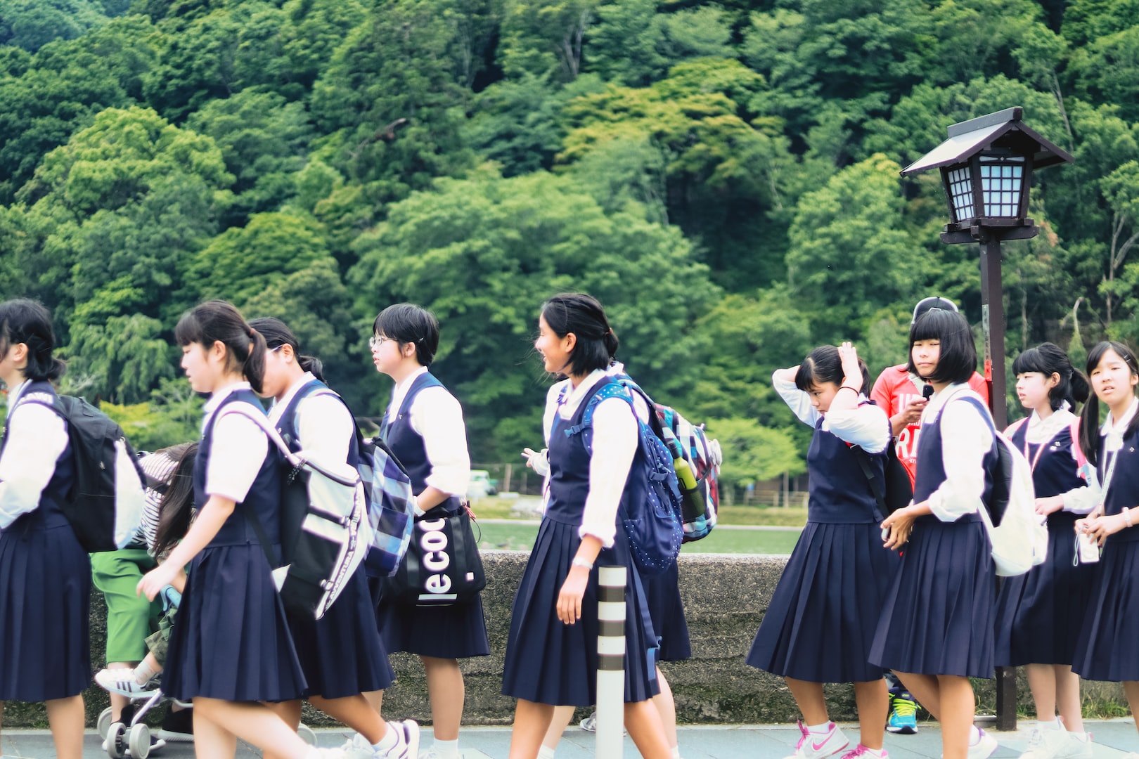 This photo features high school girls in their school uniforms. The uniforms feature a white shirt with a navy blue jumper over the shirt. The students have backpacks on their backs as well. The students are outside and behind them features an open field with many trees behind them.