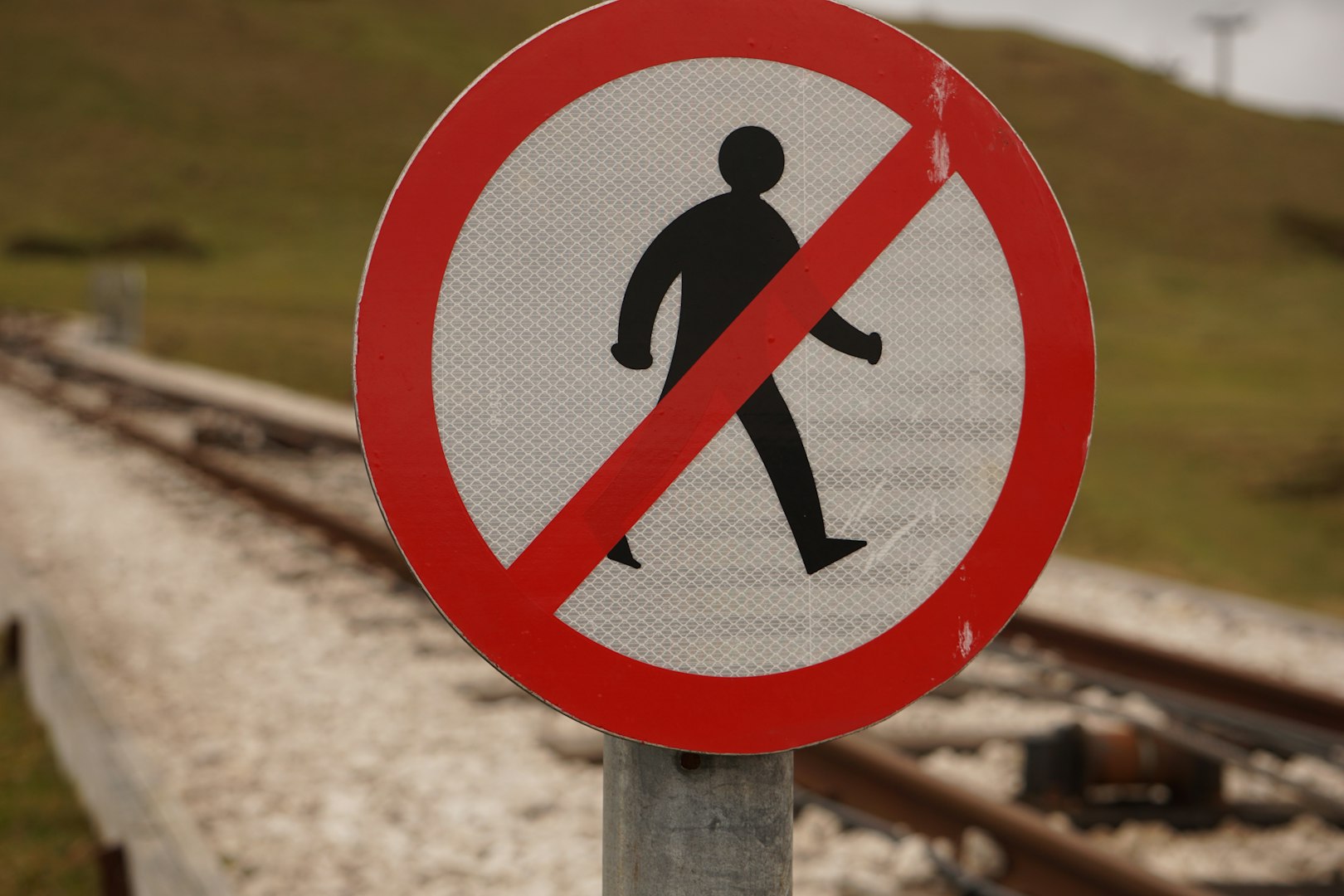 This image features a sign. The sign has a blacked out drawing of a man walking. There is a bright red circle with a line through the image of a man. This indicates restriction and that people should not walk near that area. The background is blurry, but a grassy hill can be seen. Slightly closer to the viewer is a rocky area with train tracks in the center.