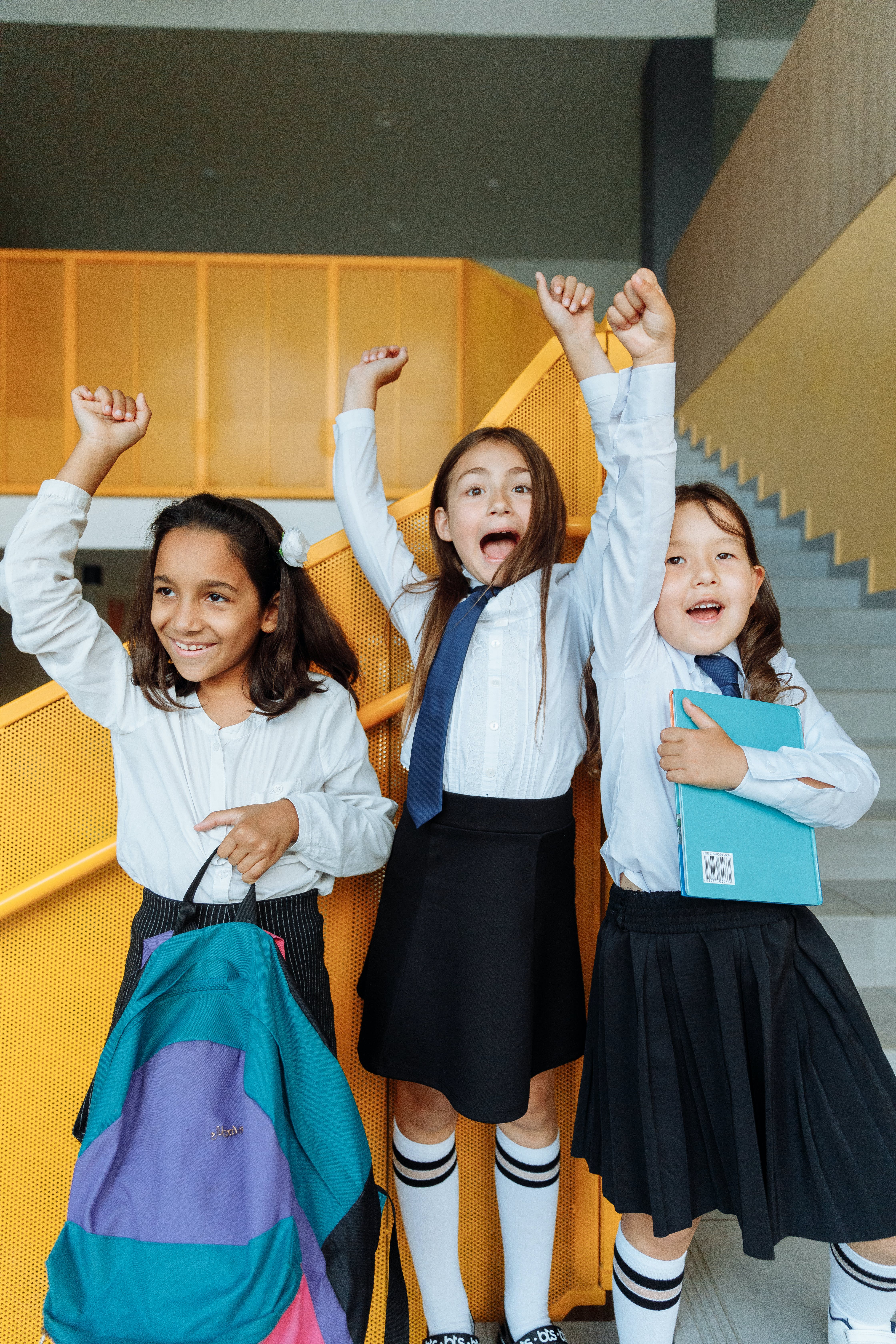 This is a photo of three young children in their school uniforms. One student is holding a backpack and another is holding what appears to be a notebook. The three students are jumping into the air and appear to be very happy.