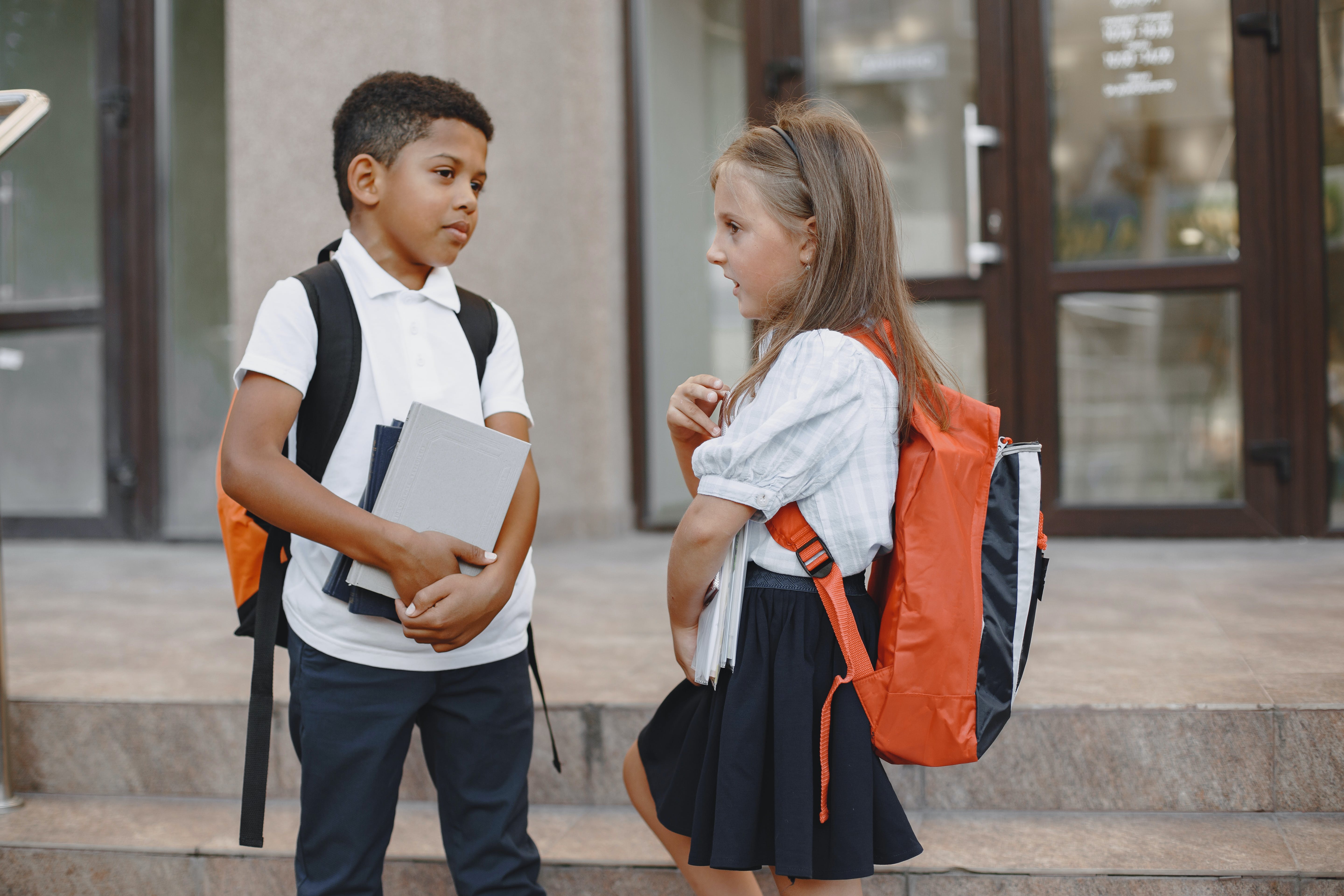 This photo features two young students, a boy and a girl. They seem to be having a conversation with each other. They both are wearing a unform. The boy is wearing a white polo and black pants. The girl is wearing a white shirt with a black skirt. They both have on their backpacks and the boy is holding paper. They are outside. They seem focused on the conversation.