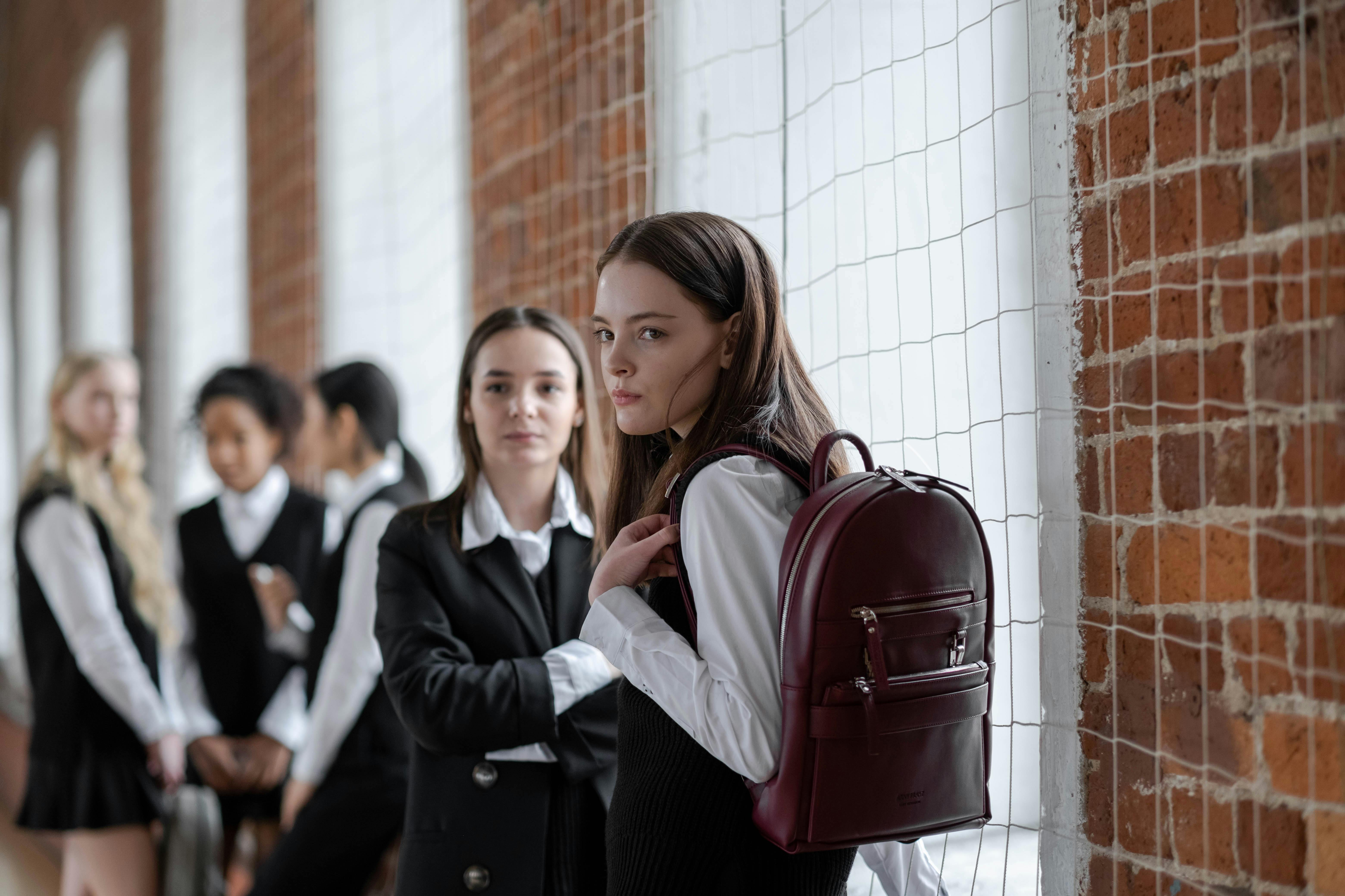 This is an image of two girls in their school uniforms. They appear to be in school with other students appearing blurry in the background. The two girls in the photo appear to look bored or disinterested.