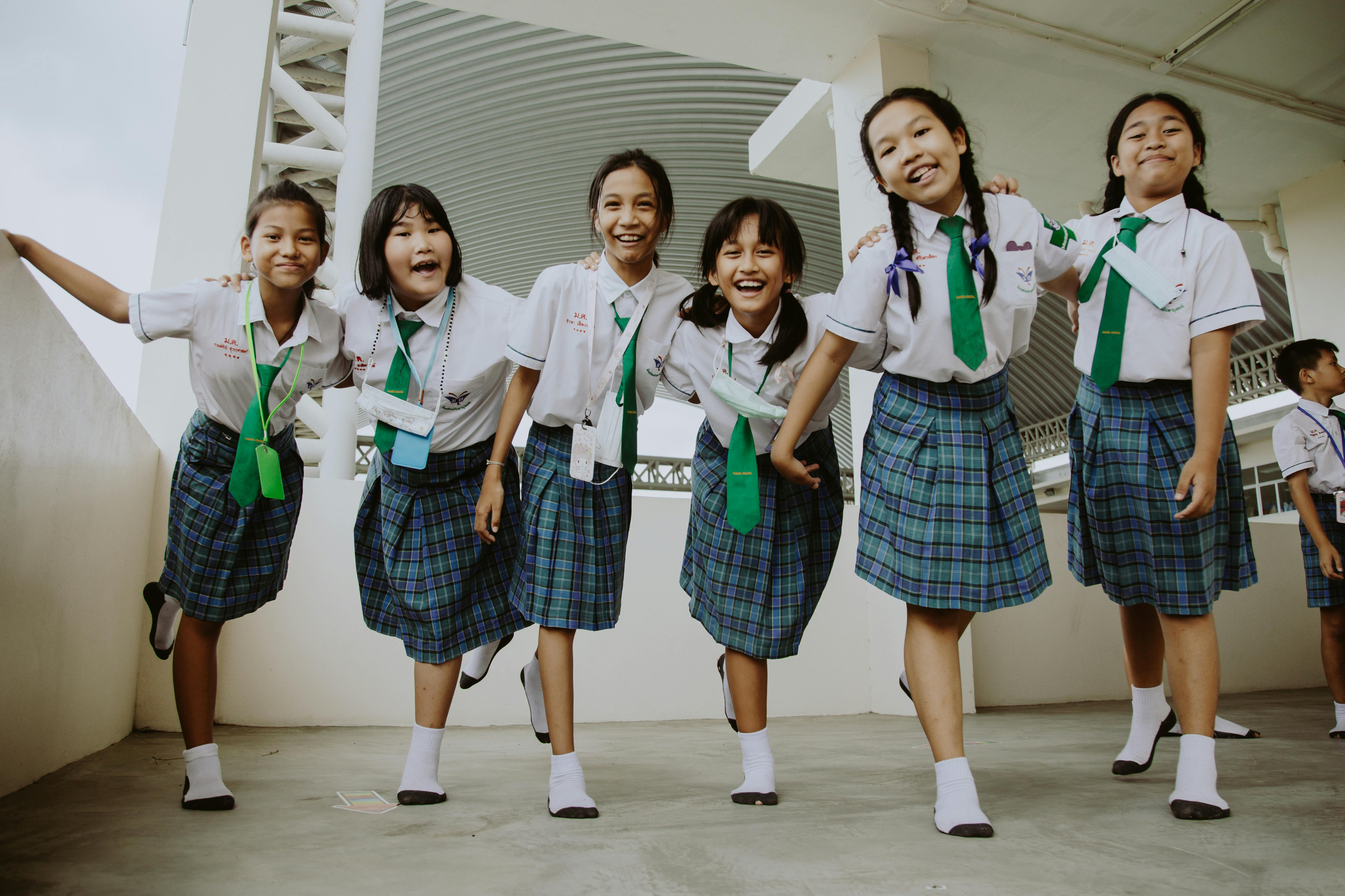 This image features six young girls all wearing the same school uniform. The uniform consists of a white button up shirt, a green tie, and a blue, green, black, and white plaid skirt. The background is not very clear as to the location the students are in. It is a white building with what appears to be concrete flooring. The students all appear to be very happy and are all smiling.